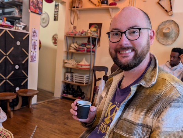 A man is sitting in a cafe holding an espresso cup and smiling at the camera.